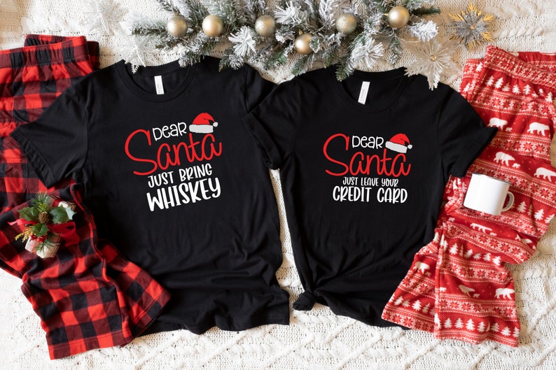 May include: Two black t-shirts with a Christmas theme. The left shirt says "Dear Santa Just Bring Whiskey" and the right shirt says "Dear Santa Just Leave Your Credit Card".