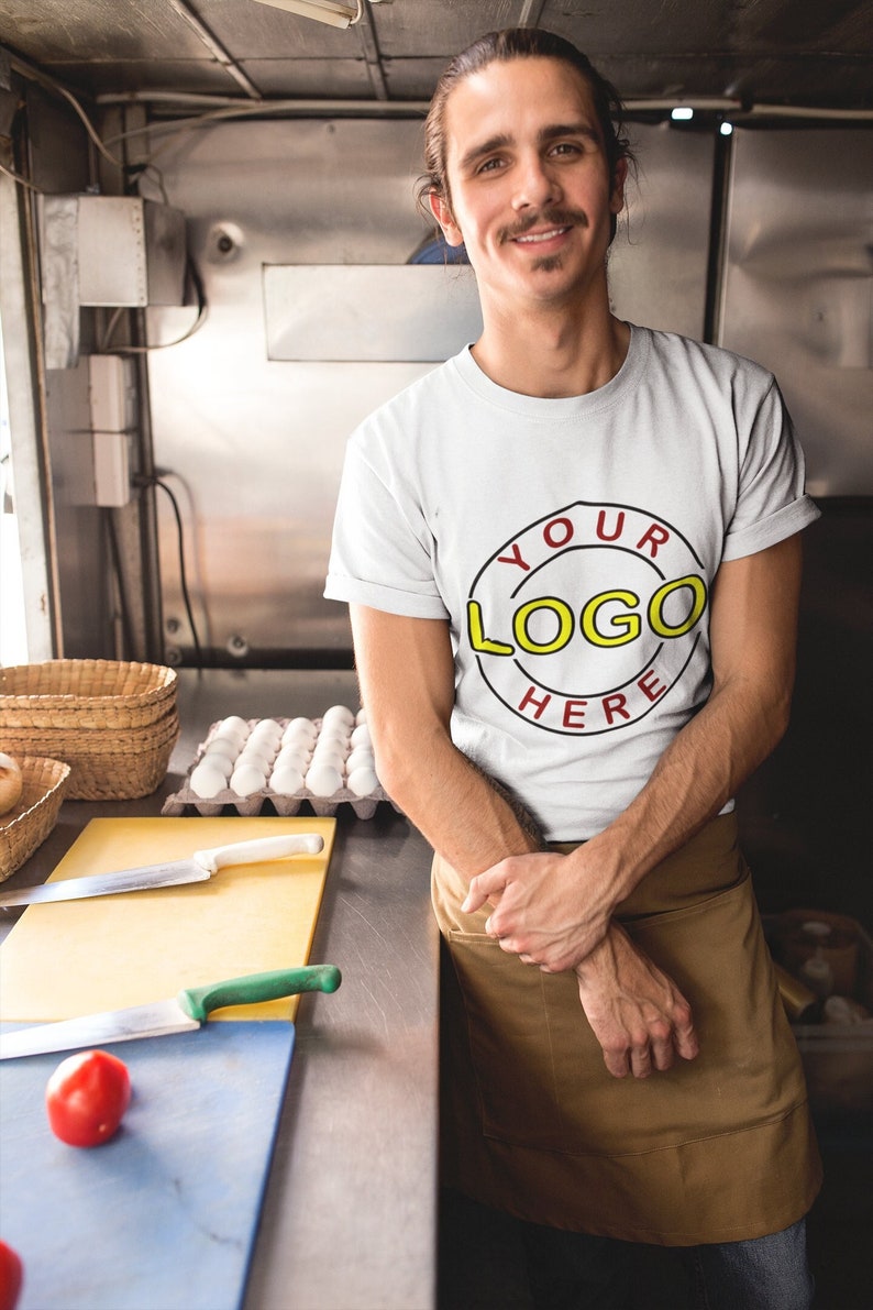 May include: A man wearing a white t-shirt with a red circle and the text "YOUR LOGO HERE" in yellow. He is standing in a kitchen with a brown apron and is smiling at the camera.