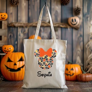 Pode incluir: Uma bolsa tote de lona bege com um design de Minnie Mouse tem&aacute;tico de Halloween. O design apresenta uma silhueta preta da cabe&ccedil;a da Minnie Mouse com um grande la&ccedil;o laranja. A silhueta est&aacute; cheia de v&aacute;rios &iacute;cones de Halloween, incluindo ab&oacute;boras, morcegos, fantasmas e teias de aranha. A bolsa tem uma al&ccedil;a preta e o nome "Sophia" est&aacute; impresso em preto abaixo do design.