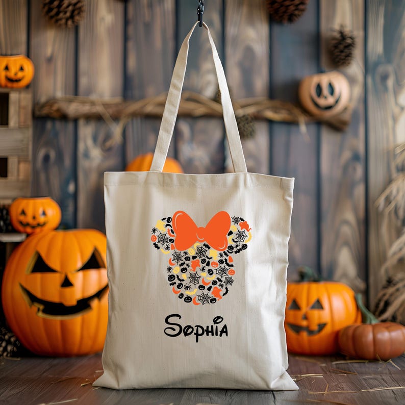 Pode incluir: Uma bolsa tote de lona bege com um design de Minnie Mouse tem&aacute;tico de Halloween. O design apresenta uma silhueta preta da cabe&ccedil;a da Minnie Mouse com um grande la&ccedil;o laranja. A silhueta est&aacute; cheia de v&aacute;rios &iacute;cones de Halloween, incluindo ab&oacute;boras, morcegos, fantasmas e teias de aranha. A bolsa tem uma al&ccedil;a preta e o nome "Sophia" est&aacute; impresso em preto abaixo do design.