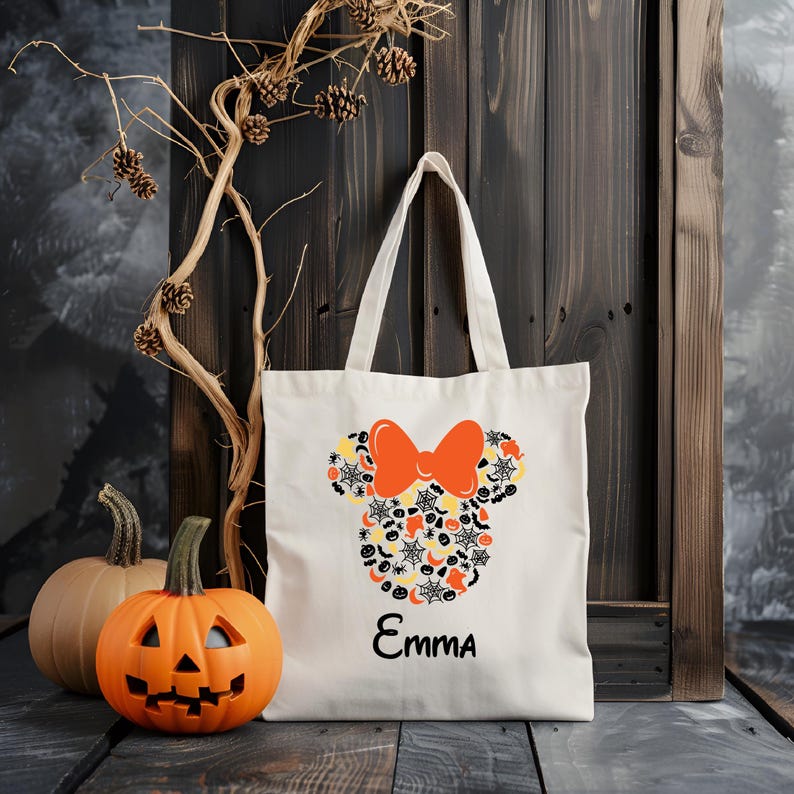 Pode incluir: Sacola de lona branca com um design de Mickey Mouse de Halloween com cores laranja, preto e amarelo. A sacola tem um la&ccedil;o preto e o nome "Emma" impresso na parte inferior.