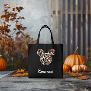 Pode incluir: Sacola de tecido preta para Halloween com um design de cabe&ccedil;a do Mickey Mouse feito de &iacute;cones de Halloween. A sacola tem uma al&ccedil;a branca e o nome "EMERSON" impresso abaixo do design.