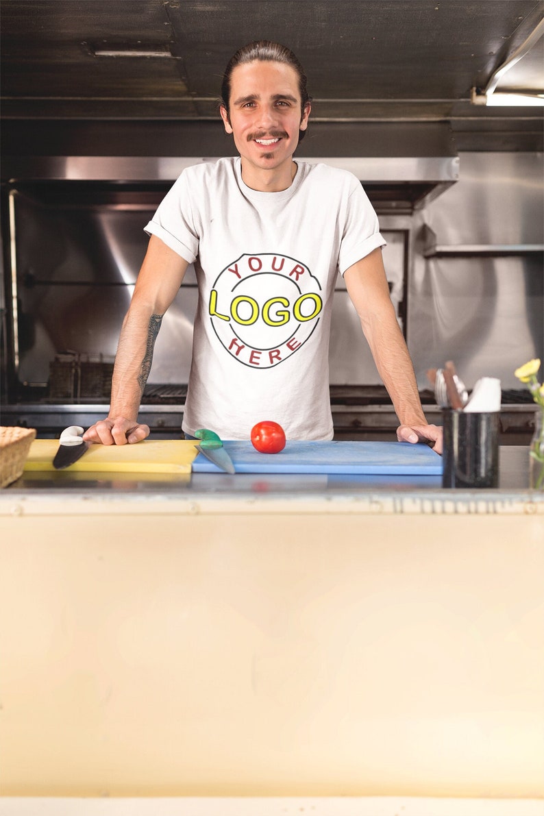 May include: A man wearing a white t-shirt with a red circle and the text "YOUR LOGO HERE" in yellow. He is standing in a kitchen with a cutting board and a tomato.