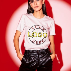 May include: A woman wearing a white t-shirt with a red circle and the text "YOUR LOGO HERE" in yellow and black. She is wearing black leather pants and has her hands in her pockets.