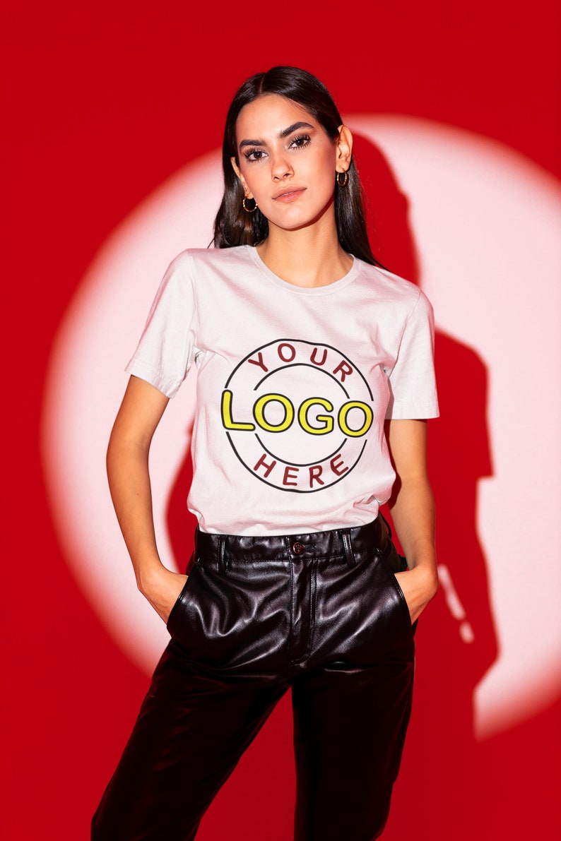May include: A woman wearing a white t-shirt with a red circle and the text "YOUR LOGO HERE" in yellow and black. She is wearing black leather pants and has her hands in her pockets.