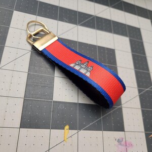 May include: A red and blue keychain wrist strap with the Cambodian flag design. It has a gold-coloured metal clasp and a black underside. The flag design includes a white temple image.