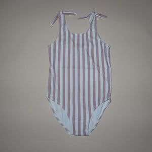 Può includere: Un costume da bagno intero a righe verticali rosa chiaro e bianco. Il costume ha spalline annodate sulle spalle e un retro a girocollo. Il fondo del costume è bianco.