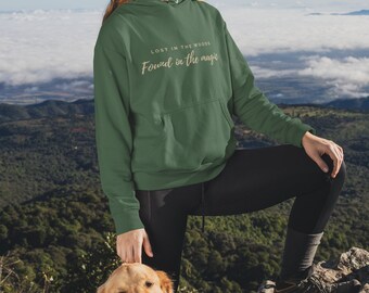 Puede incluir: Una mujer con una sudadera con capucha verde con el texto "Lost in the woods Found in the magic" está de pie en la cima de una montaña rocosa con un perro golden retriever. La mujer lleva una boina negra y gafas de sol. El fondo es un cielo azul con nubes blancas.