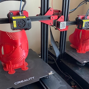Puede incluir: Dos impresoras 3D con filamento rojo imprimiendo máscaras faciales rojas. Las impresoras son ambas negras con detalles rojos y el nombre de la marca "CREALITY" es visible en la parte delantera de cada impresora.