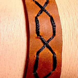 May include: A brown leather bracelet with black stitching in a criss-cross pattern.