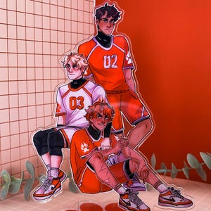 May include: Acrylic stand featuring three anime-style characters in orange and white athletic uniforms. The characters are posed together, with one seated and two standing. The stand has a paw print base and the characters wear sneakers.
