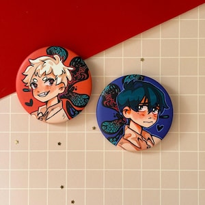 Puede incluir: Dos botones redondos con ilustraciones de estilo anime. Uno presenta un personaje con pelo rubio puntiagudo sobre un fondo rojo, el otro tiene un personaje con pelo azul oscuro sobre un fondo azul. Ambos tienen un corazón.