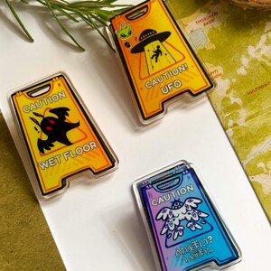 May include: Three novelty caution sign pins. One yellow pin reads "CAUTION! UFO" with an alien and a person being beamed up. Another reads "WET FLOOR" with a monster. The third pin is blue and purple with Japanese text.
