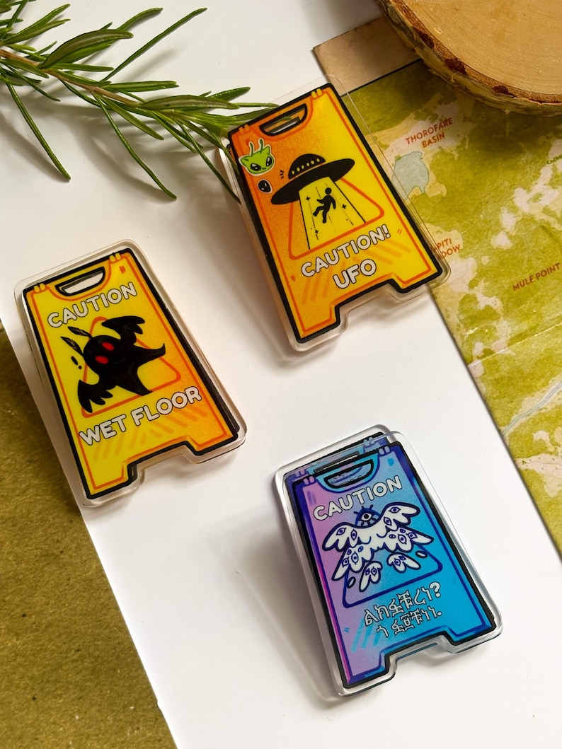 May include: Three novelty caution sign pins. One yellow pin reads "CAUTION! UFO" with an alien and a person being beamed up. Another reads "WET FLOOR" with a monster. The third pin is blue and purple with Japanese text.