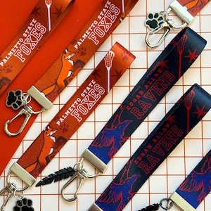 Può includere: Diversi cordini con loghi e mascotte di squadre. I cordini Palmetto State Foxes sono arancioni con una volpe e un bastone da lacrosse. I cordini Edgar Allan Ravens sono blu navy con un corvo. Ogni cordino ha una chiusura in metallo.