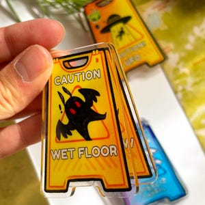 May include: A set of novelty acrylic signs with a yellow and black caution design. The sign reads "CAUTION WET FLOOR" with a black creature graphic. Additional signs feature UFO themes. The signs are held in a hand.