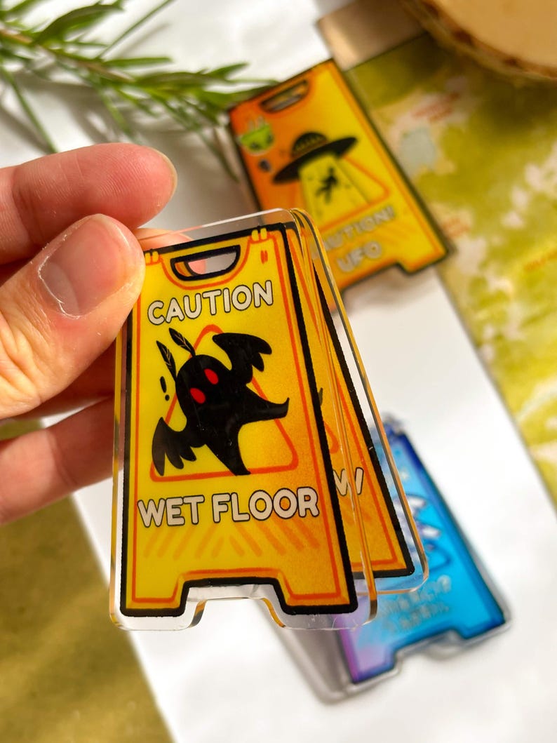 May include: A set of novelty acrylic signs with a yellow and black caution design. The sign reads "CAUTION WET FLOOR" with a black creature graphic. Additional signs feature UFO themes. The signs are held in a hand.
