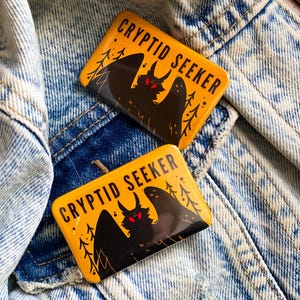 Op de afbeelding: Twee rechthoekige gele pins met de tekst "CRYPTID SEEKER" en een zwarte illustratie van een wezen met rode ogen en vleugels. De pins liggen op een spijkerjasje.