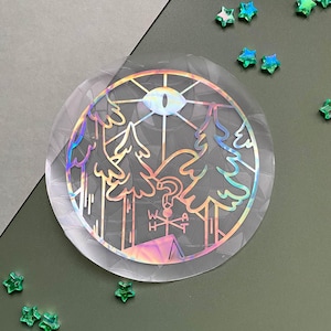 Puede incluir: Una pegatina holográfica circular con un diseño de árboles, un ojo y un signo de interrogación. La pegatina tiene un brillo de arcoíris y un borde transparente. Adornos verdes en forma de estrella están dispersos alrededor.
