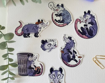 May include: A set of nine stickers featuring cartoon opossums in various poses and expressions. The stickers are printed on white paper and have a glossy finish. The opossums are depicted in a variety of colors, including purple, pink, and blue. Some of the stickers feature text, such as "Crime!" and "a?"
