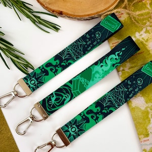 May include: Three green and black patterned lanyards with silver metal clasps. The lanyards display designs such as spiderwebs, geometric shapes, and cassette tapes. The background includes a wooden slice, rosemary sprigs, and a map.