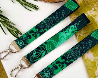 Può includere: Tre cordini verdi e neri a fantasia con chiusure in metallo color argento. I cordini presentano vari disegni, tra cui ragnatele, forme geometriche e musicassette. Lo sfondo comprende una fetta di legno, rametti di rosmarino e una mappa.