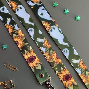 May include: Three lanyards with a repeating pattern of light blue ghosts and sunflowers on a dark green background. Each lanyard has a metal clasp and a green leather end. Scattered around are green star-shaped embellishments and a gold paperclip.