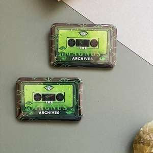 May include: Two green and black cassette tape-shaped pins with the text "The Magnus Archives" on them. The pins have a green eye design around the edges.