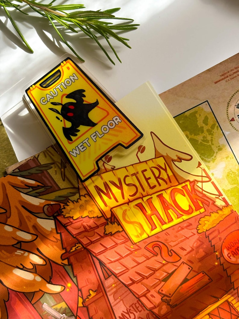 May include: A yellow "Caution Wet Floor" sign with a black silhouette of a creature. The sign is placed on top of a book with the words "Mystery Shack" in orange. A sprig of rosemary is in the background.