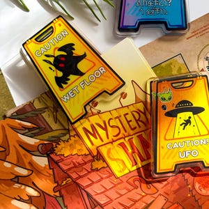 May include: A collection of colorful, cartoon-style acrylic pins. One pin features a yellow caution sign with a black creature and the words "WET FLOOR." Another has a UFO theme with the text "CAUTION! UFO."