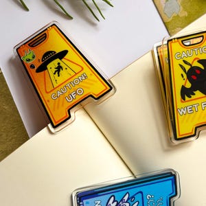 May include: Acrylic pins with a yellow caution sign design. One pin features a UFO and alien graphic with the text "CAUTION! UFO". Another pin has a black creature graphic. The third pin has a blue and purple design with text.