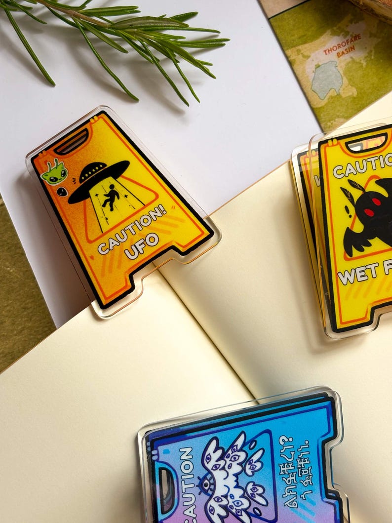 May include: Acrylic pins with a yellow caution sign design. One pin features a UFO and alien graphic with the text "CAUTION! UFO". Another pin has a black creature graphic. The third pin has a blue and purple design with text.