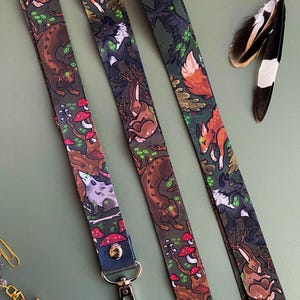 May include: Three green, black, and brown patterned lanyards with a gold metal clip. The lanyards feature a repeating pattern of foxes, rabbits, owls, and mushrooms.
