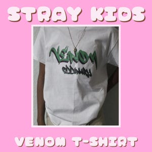 Puede incluir: Camiseta blanca con un estampado gráfico verde que dice "Venom" y "Oddinary" en estilo graffiti. La camiseta tiene una cadena de oro en la parte superior.