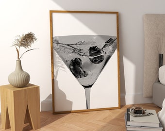 May include: A black and white photograph of a martini glass with olives in it, framed in a wooden frame and hanging on a white wall above a wooden side table.