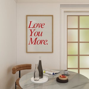 May include: A framed print with the words "Love You More" in red lettering hangs on a white wall. A round table with a marble top holds bottles, a tray, and a cup of coffee. A chair with a curved back is on the left.