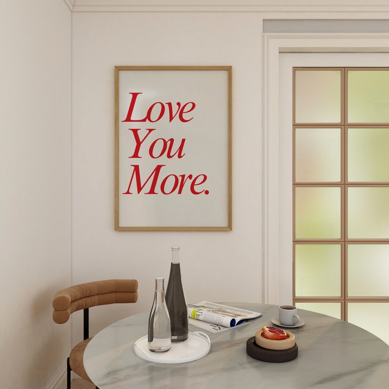 May include: A framed print with the words "Love You More" in red lettering hangs on a white wall. A round table with a marble top holds bottles, a tray, and a cup of coffee. A chair with a curved back is on the left.
