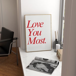 以下が含まれることがあります： 赤い文字で「Love You Most.」と書かれた白い額縁のポスター。ポスターは、雑誌とろうそくの隣にある白い大理石のカウンタートップの上に置かれています。