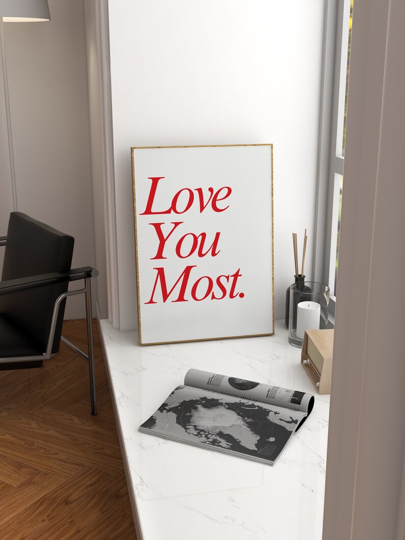 以下が含まれることがあります： 赤い文字で「Love You Most.」と書かれた白い額縁のポスター。ポスターは、雑誌とろうそくの隣にある白い大理石のカウンタートップの上に置かれています。
