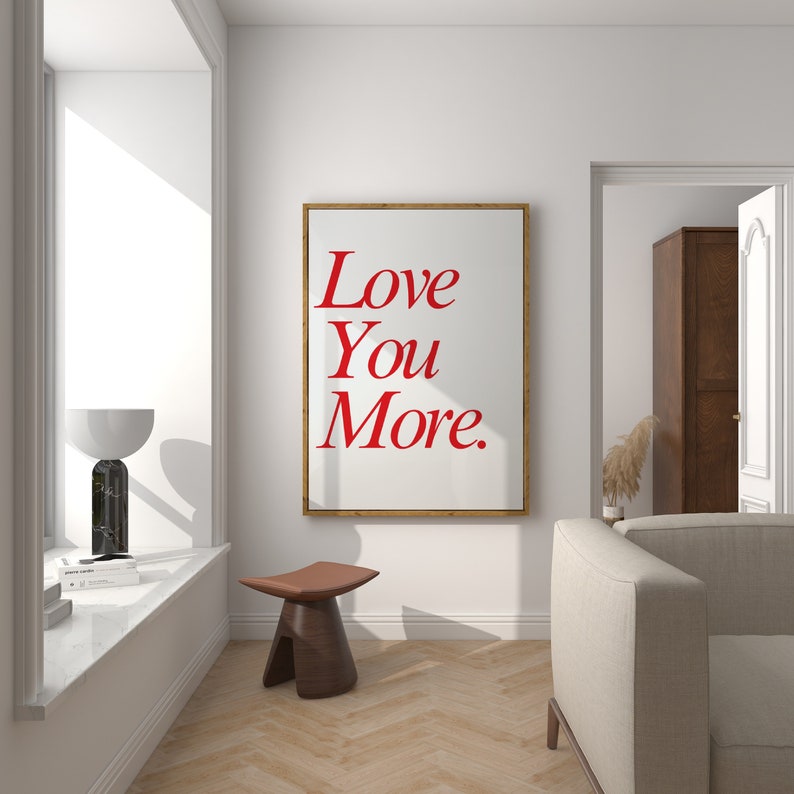 May include: A framed art print with the words "Love You More" in red script. The print is displayed on a white wall in a room with a window, a wooden stool, and a sofa. The frame is a light brown color.