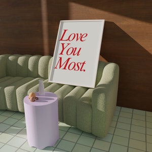 以下が含まれることがあります： 赤い文字で「Love You Most」と書かれた白い額縁のプリントが、薄い紫色のサイドテーブルのある緑色のソファに寄りかかっています。