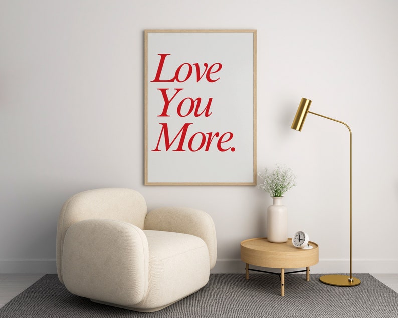May include: A framed print with the words "Love You More" in red script. The print hangs above a beige armchair, a small wooden side table, and a gold floor lamp. A vase of white flowers and a small clock are on the table.