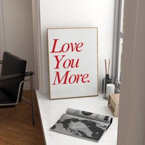 May include: A framed art print with the words "Love You More" in red script on a white background. The print is displayed on a white windowsill, alongside a magazine, candle, and a small radio.
