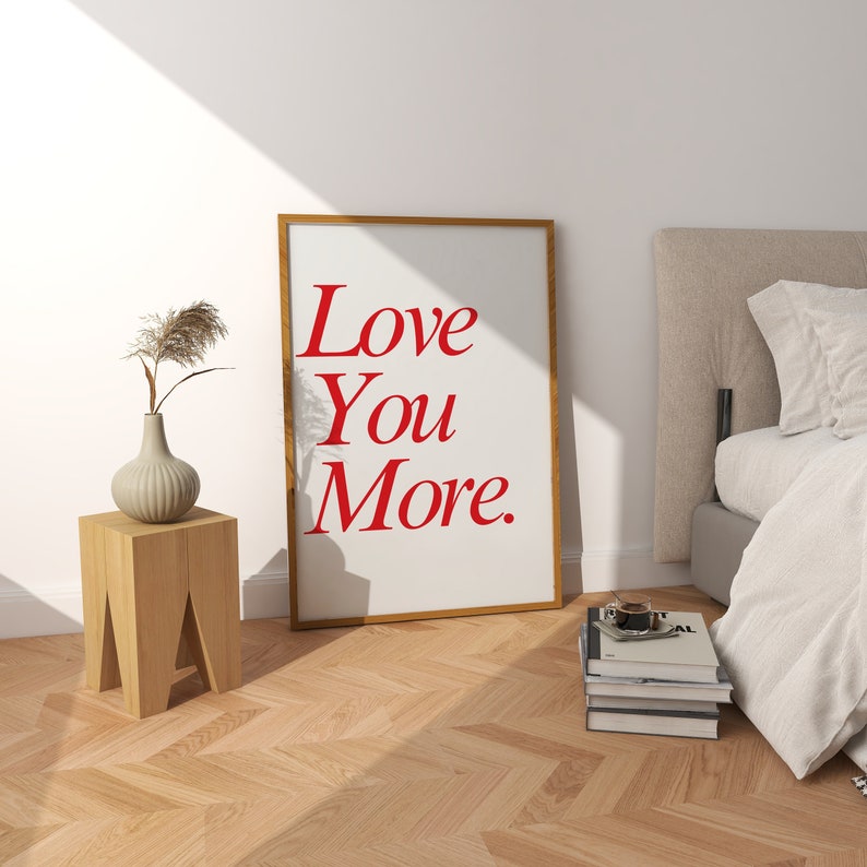 May include: A framed art print with the words "Love You More" in red lettering. The print leans against a wall near a bed and a small wooden side table with a vase and decorative plant. The room has a light, airy feel.