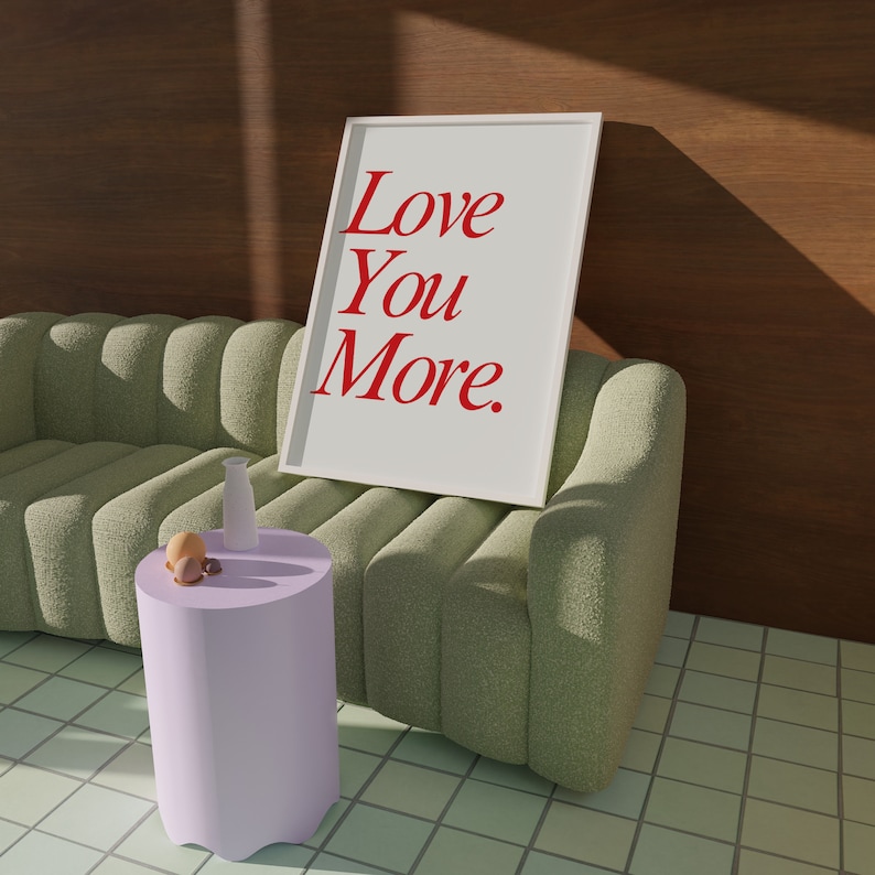 May include: A framed print with the words "Love You More" in red script. The print leans against a light green, textured sofa. A small, lavender side table holds a white vase and decorative objects. The background features a wooden wall.