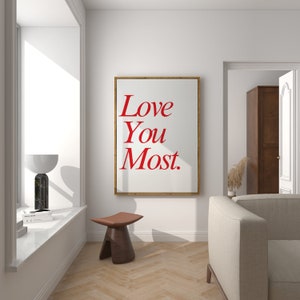 以下が含まれることがあります： 白い背景に赤い文字で「Love You Most.」と書かれた額装されたプリント。プリントは、木製の床と窓のある部屋の白い壁にかけられています。