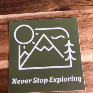 Può includere: Adesivo verde con un contorno bianco di una catena montuosa, un sole e un albero all'interno di una cornice. Il testo "Never Stop Exploring" è sotto l'immagine.