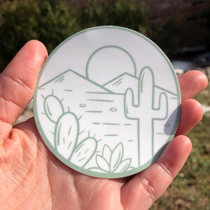 Puede incluir: Una pegatina circular blanca con un contorno verde presenta un dibujo lineal minimalista de un paisaje desértico con un cactus, montañas y un sol.