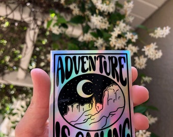 Könnte beinhalten: Ein holografischer Aufkleber mit einem Schwarz-Weiß-Design eines Halbmondes und einer Bergkette. Der Text "Adventure is Calling" ist in einer stilisierten Schriftart geschrieben.