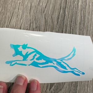 May include: A bright blue vinyl decal featuring a stylized silhouette of a dog in a running or leaping pose with its mouth open. The decal is presented on a white rectangular backing paper, showing a dynamic canine shape with sharp lines.